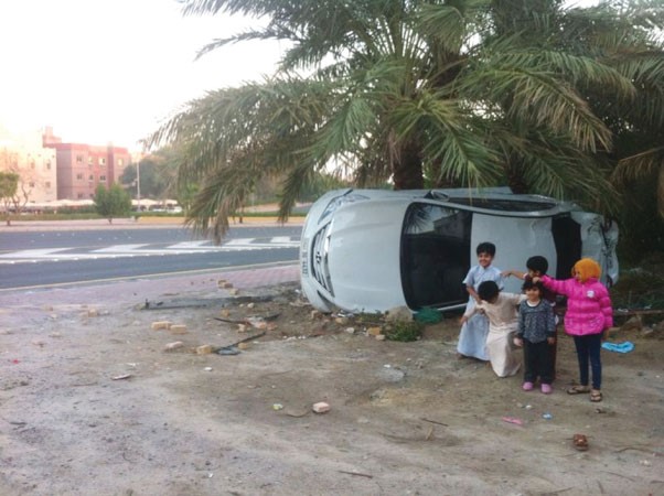 اطفال بجانب السيارة المنحرفة بعد الحادث ويبدو المنعطف الخطير المواجه لمنزل العجمي ﻿