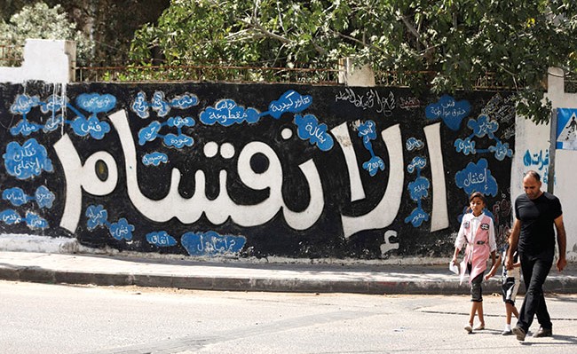 فلسطيني وابنته يمران امس بجوار جدارية تشير الى الانقسام بين حركتي «فتح» و«حماس» وتداعياته السلبية (أ.ف.پ)