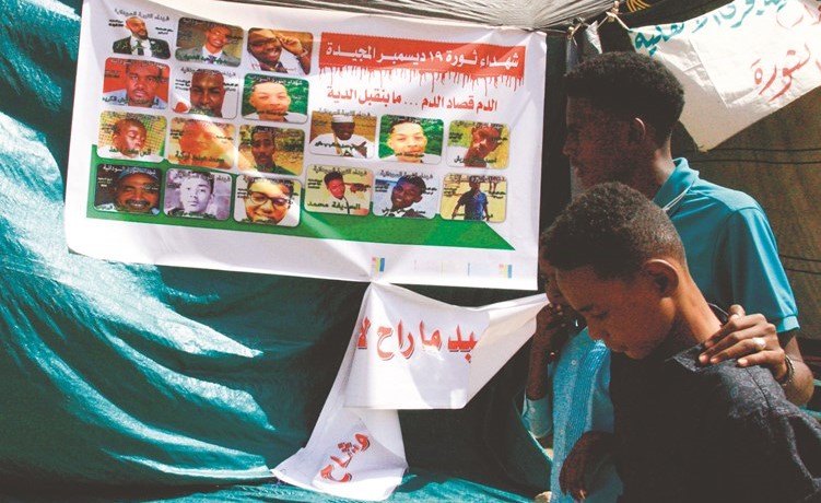 سودانيون يسيرون بجانب لافتة تحمل صور المتظاهرين الذين لقوا حتفهم خلال الاحتجاجات خارج مقر الجيش في الخرطوم أمس (أ.ف.پ)﻿